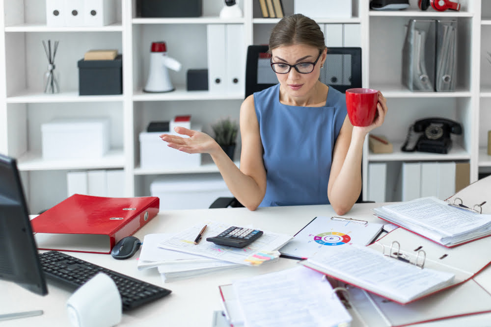 Woman is sitting at a table in the office, holding a red cup in her hand and working with many documents.